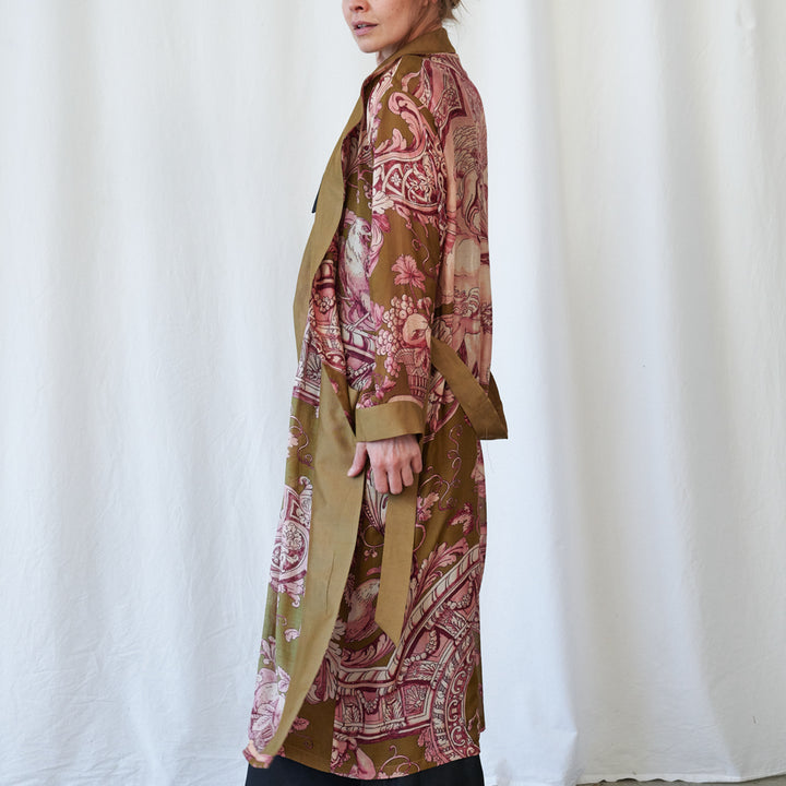 A model shown from the side wearing a long-sleeved, open-front kimono-style robe with a brown base and pink paisley pattern, set against a white draped fabric backdrop.