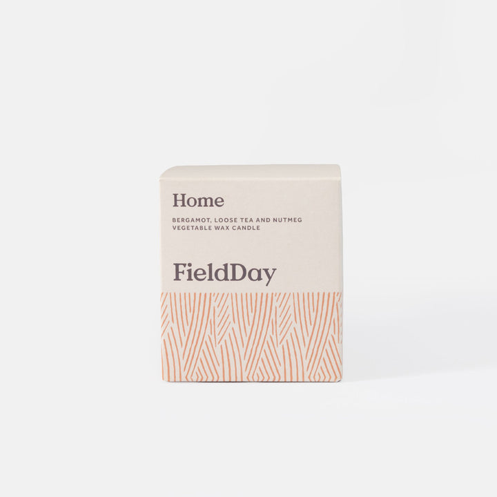 A square FieldDay 'Home' vegetable wax candle box, featuring a light beige top half with dark gray text and a bottom half with a terracotta orange pattern of wavy vertical lines, set against a clean white background.