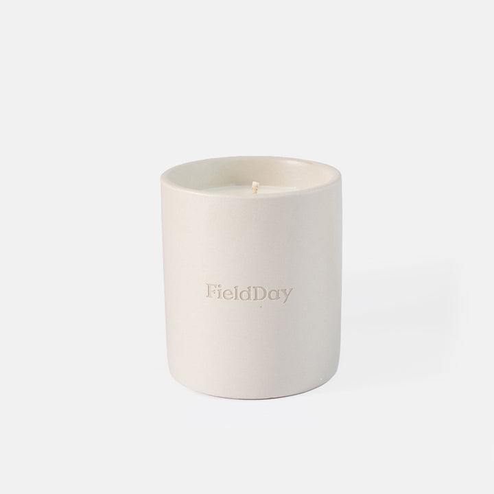 A minimalist ceramic candle in a matte off-white container with the brand name 'FieldDay' embossed on the front, set against a seamless light gray background.