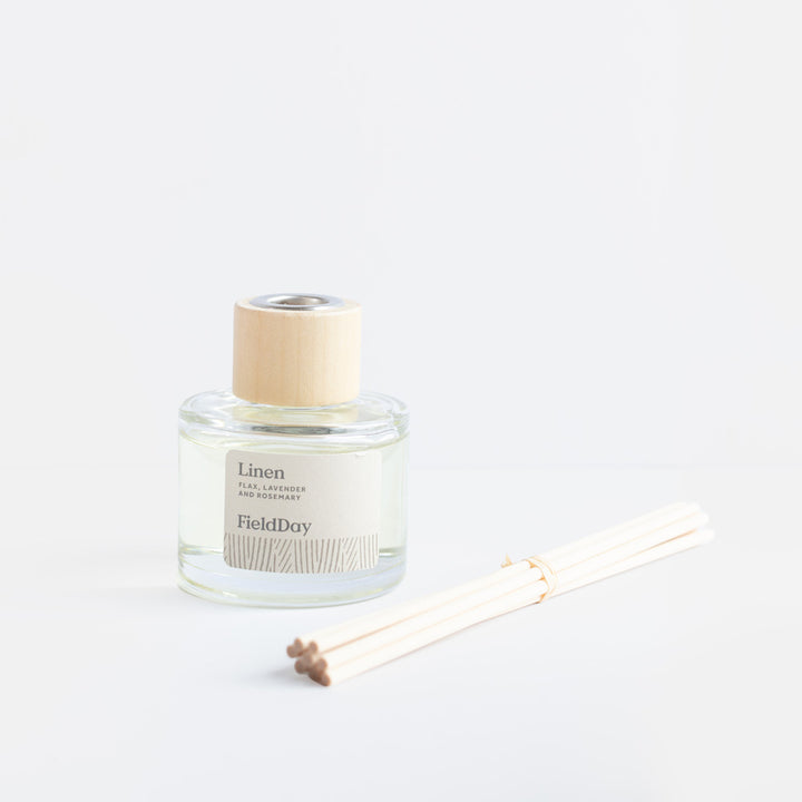 A clear glass reed diffuser bottle labeled 'Linen' and 'FieldDoy' with a light wood cap, shown next to several natural reed sticks on a clean white background.