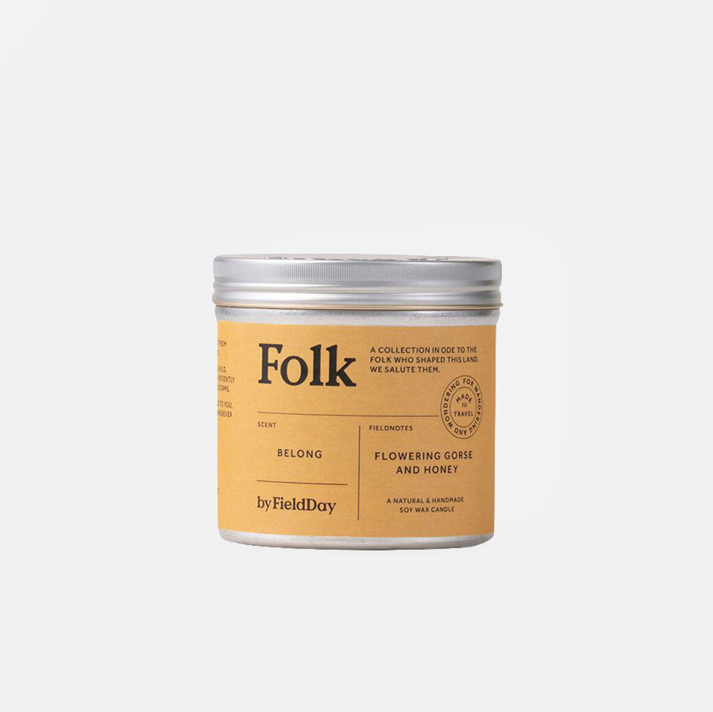 A natural wax candle in a cylindrical metal tin with a silver lid and a warm tan label. The label features the brand name 'Folk' and scent name 'Belong' by FieldDay.