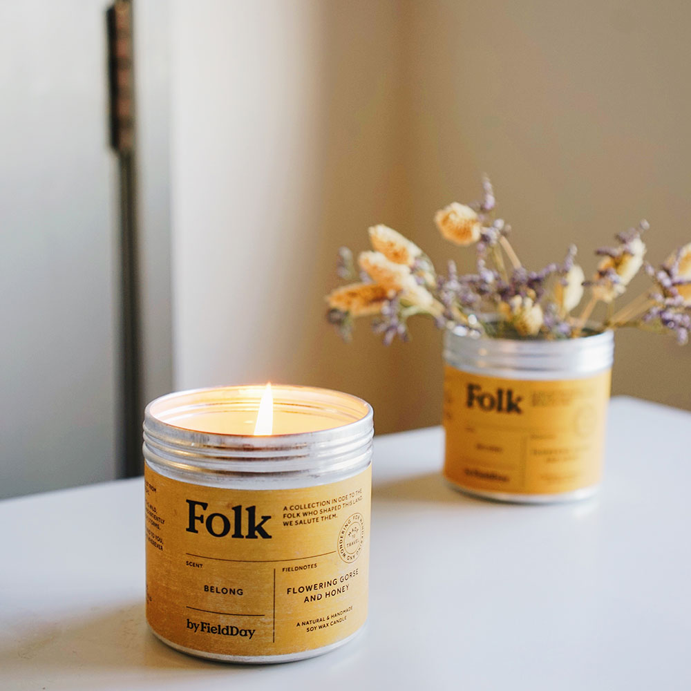 A lit Folk candle in a silver metal tin with a yellow label sits on a white tabletop, with a second matching candle and dried flowers blurred in the background.