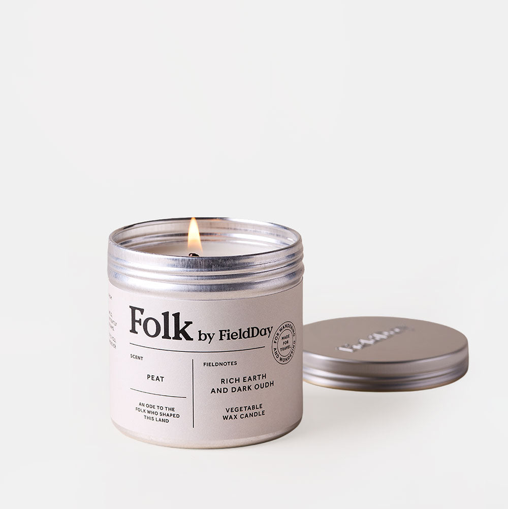A lit Folk by FieldDay candle in a silver metal tin with a white label, shown next to its matching silver lid on a clean white background.