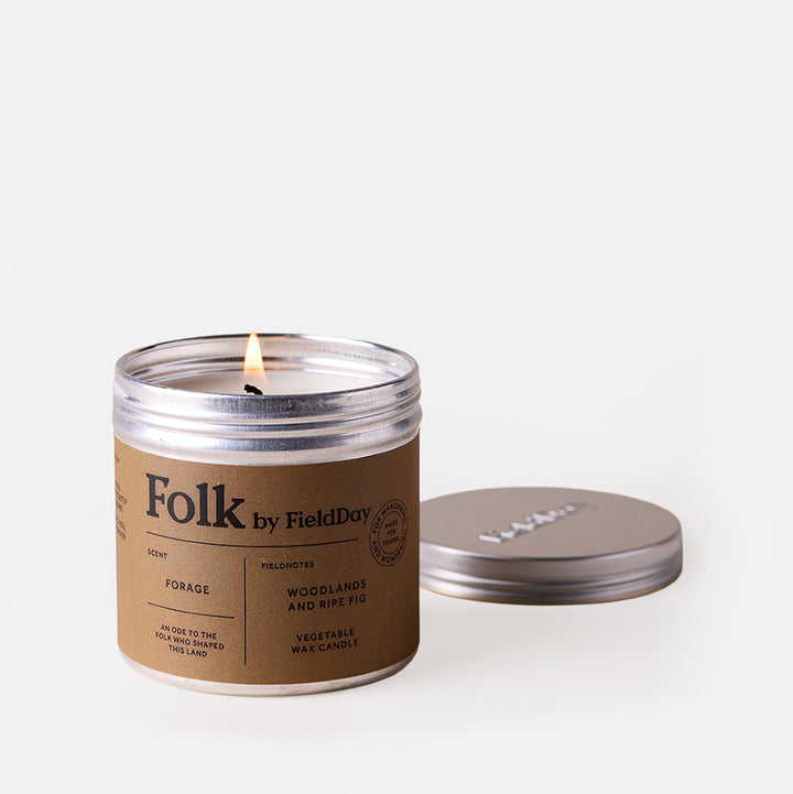 A lit candle in a metal jar with a kraft brown paper label reading 'Folk by FieldDay', shown next to its silver metal lid on a clean white background.