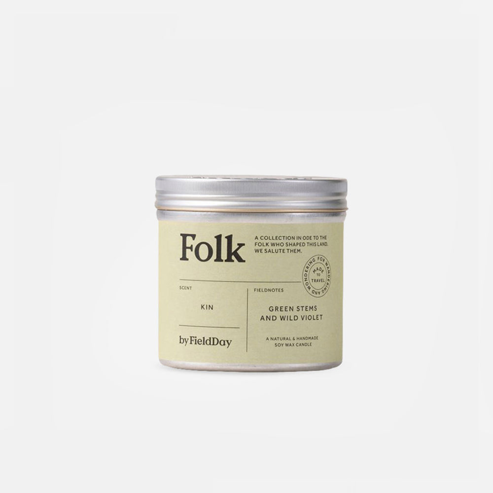 A cylindrical metal tin candle with a silver lid and a cream-colored label. The label features the brand name 'Folk' and the scent 'Kin', with 'Green Stems and Wild Violet' written below, set against a plain light gray background.