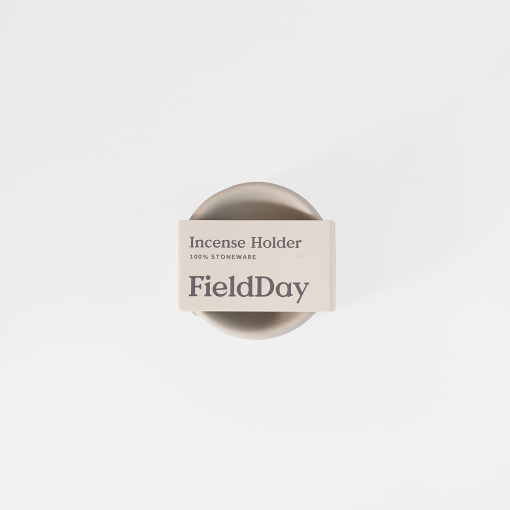 A top-down view of a FieldDay stoneware incense holder, featuring a round beige ceramic base with a white paper band label wrapped around it, set against a clean white background.