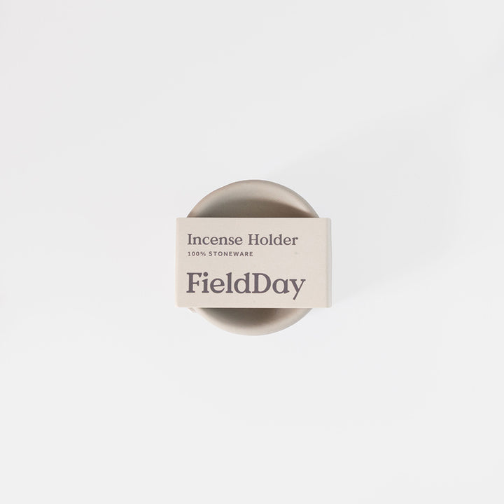 A top-down view of a FieldDay stoneware incense holder, featuring a round beige ceramic base with a white paper band label wrapped around it, set against a clean white background.