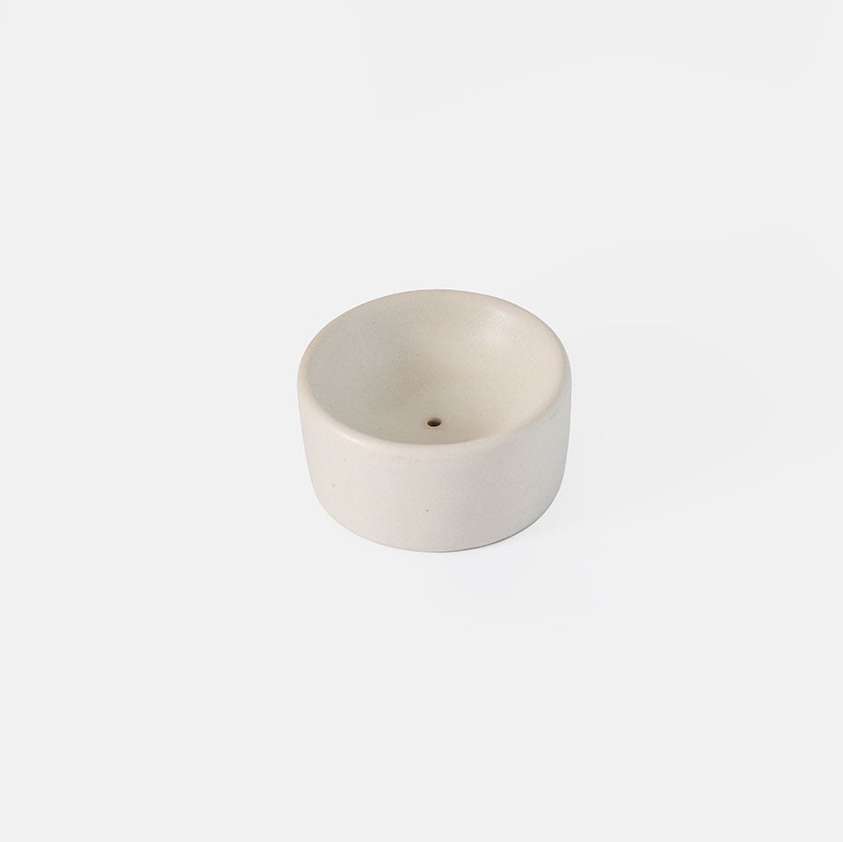 A small, round, off-white stoneware incense holder with a single central hole, shown on a seamless white background.
