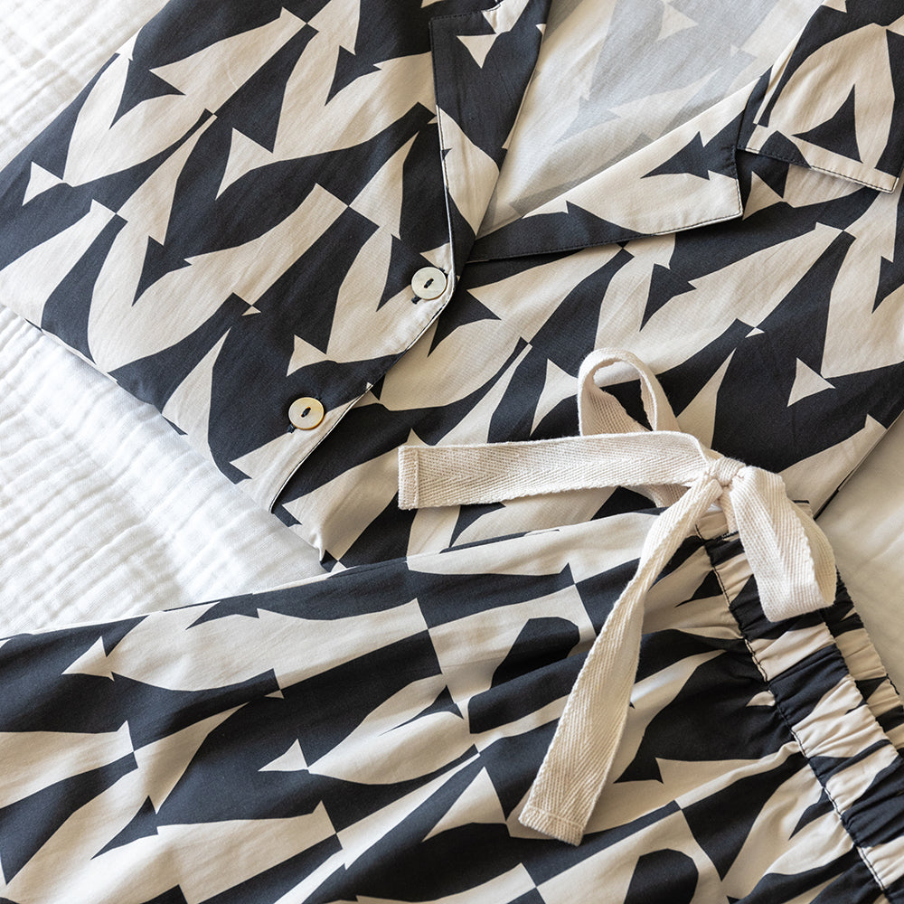 A two-piece women's pyjama set featuring a long-sleeve button-up shirt, wide-leg pants, all in a black and cream geometric hearts pattern laid out on a bed