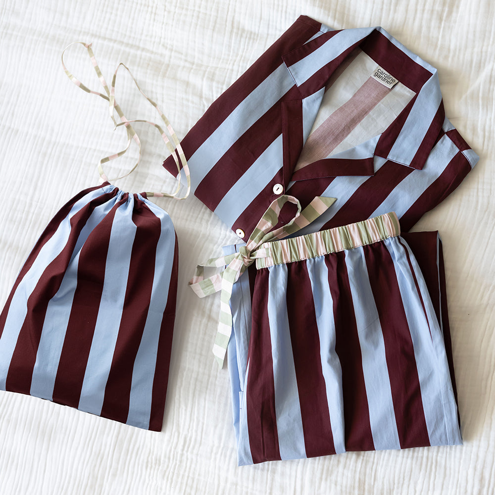 Folded blue and wine striped pyjama set with a striped tie waist, beside a matching drawstring bag on white bedding