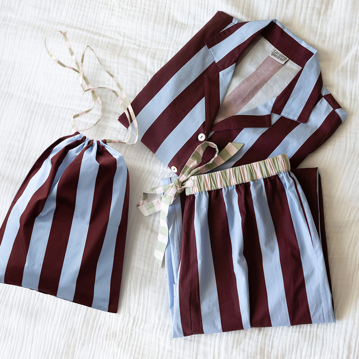 Folded blue and wine striped pyjama set with a striped tie waist, beside a matching drawstring bag on white bedding