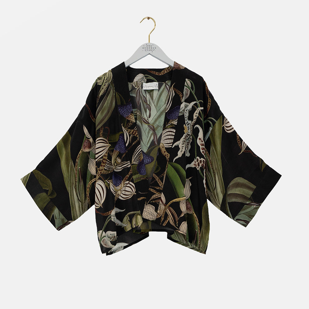 A black kimono-style blouse with wide three-quarter sleeves, featuring a detailed botanical and insect print in olive green, beige, white, and purple. The blouse is displayed on a white hanger with a gold hook against a light gray background.