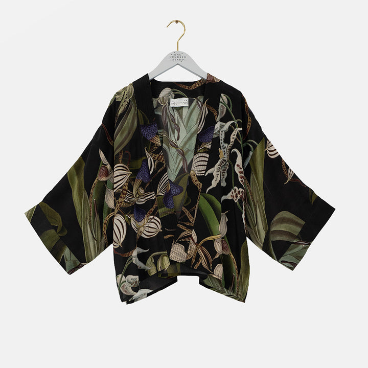 A black kimono-style blouse with wide three-quarter sleeves, featuring a detailed botanical and insect print in olive green, beige, white, and purple. The blouse is displayed on a white hanger with a gold hook against a light gray background.