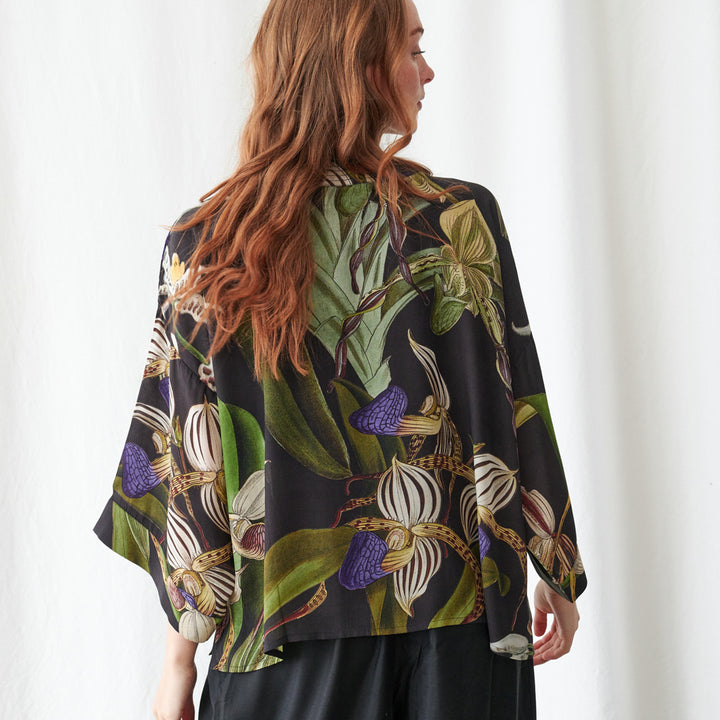 A woman shown from the back wearing a loose, wide-sleeved black blouse with a large botanical orchid print in green, purple, white, and gold tones, paired with black pants against a white curtain background.