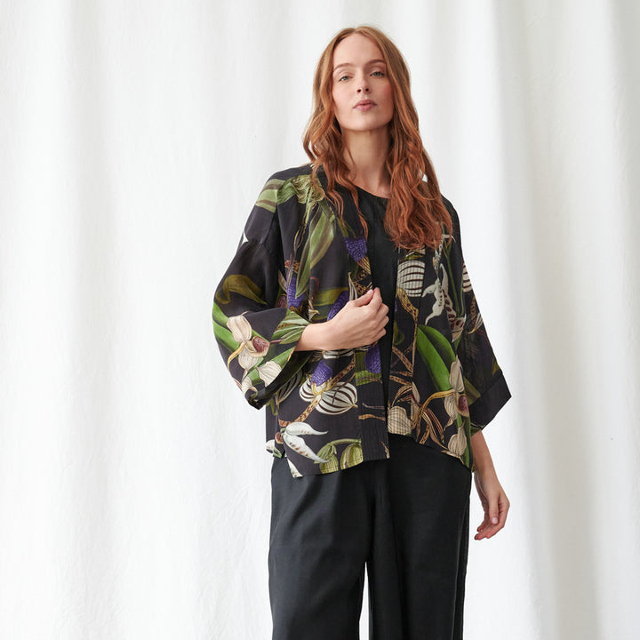 A woman modeling a black wide-sleeve kimono-style jacket with a vibrant tropical leaf and floral print in green, purple, and beige, paired with black wide-leg trousers against a white curtain backdrop.