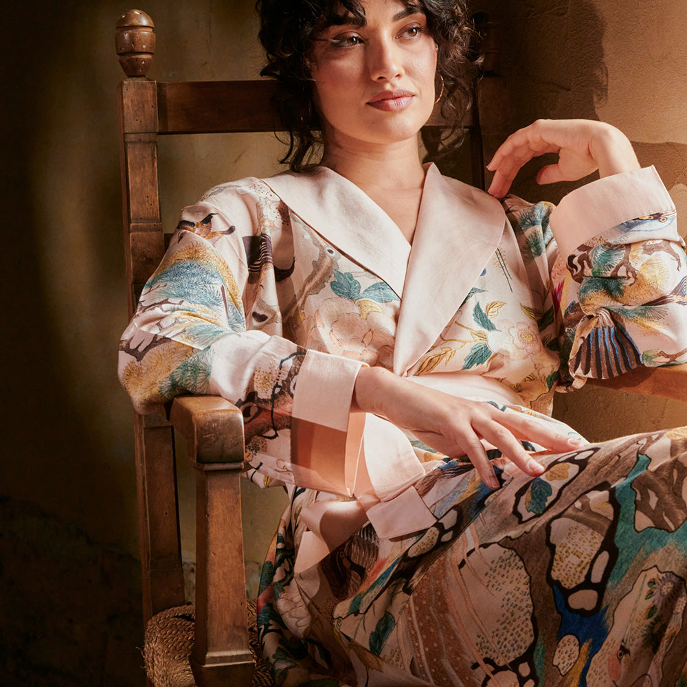 A woman sitting in a wooden chair wearing a nude chinoiserie print dressing gown with a blush pink shawl collar and cuffs.