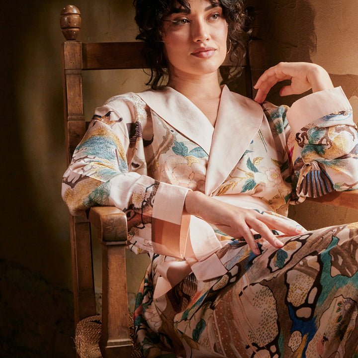 A woman sitting in a wooden chair wearing a nude chinoiserie print dressing gown with a blush pink shawl collar and cuffs.