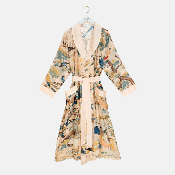 A long nude dressing gown with a hummingbird, branch and blossom print in teal, blue and ochre, with a blush pink shawl collar, cuffs, pocket trims and tie belt, on a white hanger against a light grey background.
