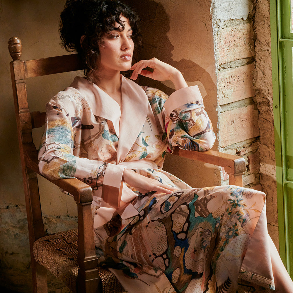 A woman seated by a window wearing a nude hummingbird print dressing gown with blush pink collar and cuffs.