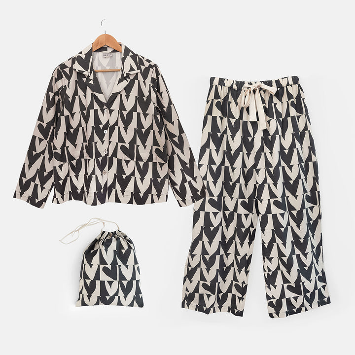 A two-piece women's pyjama set featuring a long-sleeve button-up shirt, wide-leg pants, and a drawstring storage bag, all in a black and cream geometric hearts pattern, displayed on a clean white background.