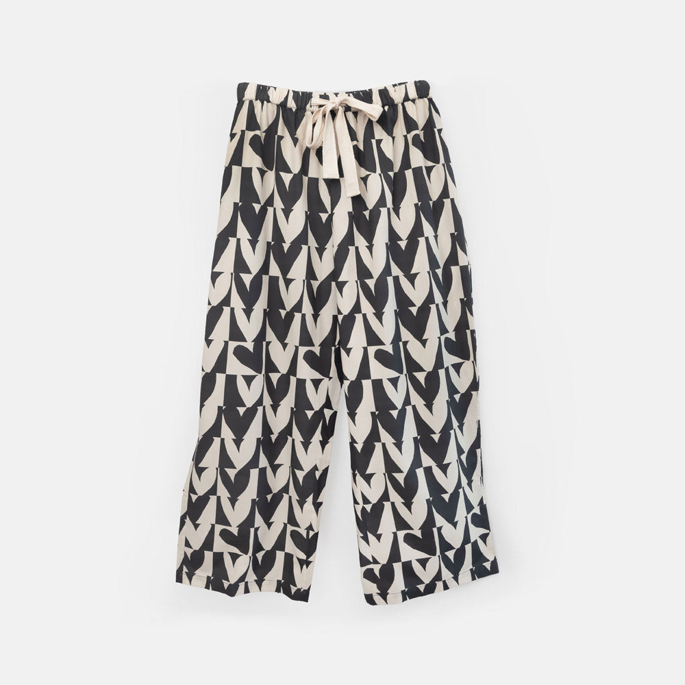 A pair of wide-leg cropped pyjama pants featuring a black and cream repeating geometric heart pattern, with a cream drawstring tie at the elastic waistband, shown against a plain light gray background.