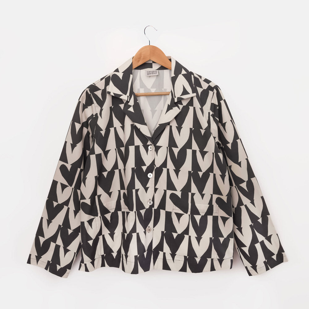 A women's long-sleeve button-up pyjama shirt with a bold black and beige geometric heart pattern, hanging on a wooden hanger against a plain white background.