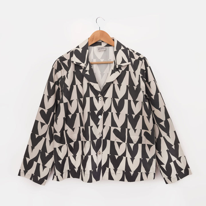 A women's long-sleeve button-up pyjama shirt with a bold black and beige geometric heart pattern, hanging on a wooden hanger against a plain white background.