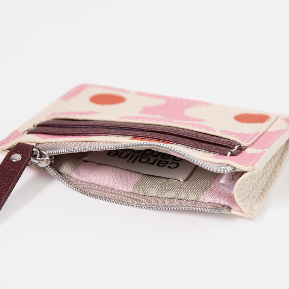A close-up of an open Caroline Gardner zip-around card holder wallet. The exterior features a light pink and cream pattern with orange-red dots, a dark brown leather wristlet strap, and silver-tone zippers.