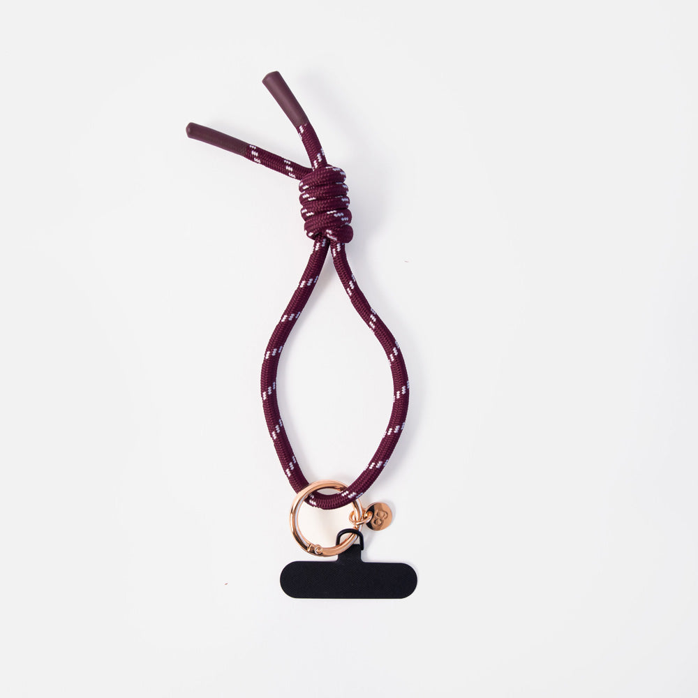A dark burgundy rope wristlet phone strap with a white speckled pattern, featuring a knot, a rose gold ring, and a black blank tag.