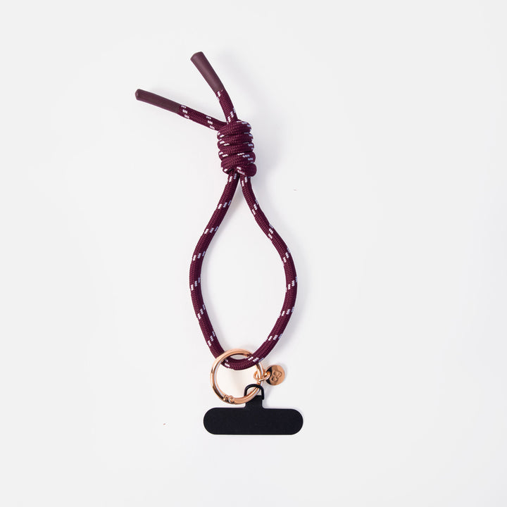 A dark burgundy rope wristlet phone strap with a white speckled pattern, featuring a knot, a rose gold ring, and a black blank tag.