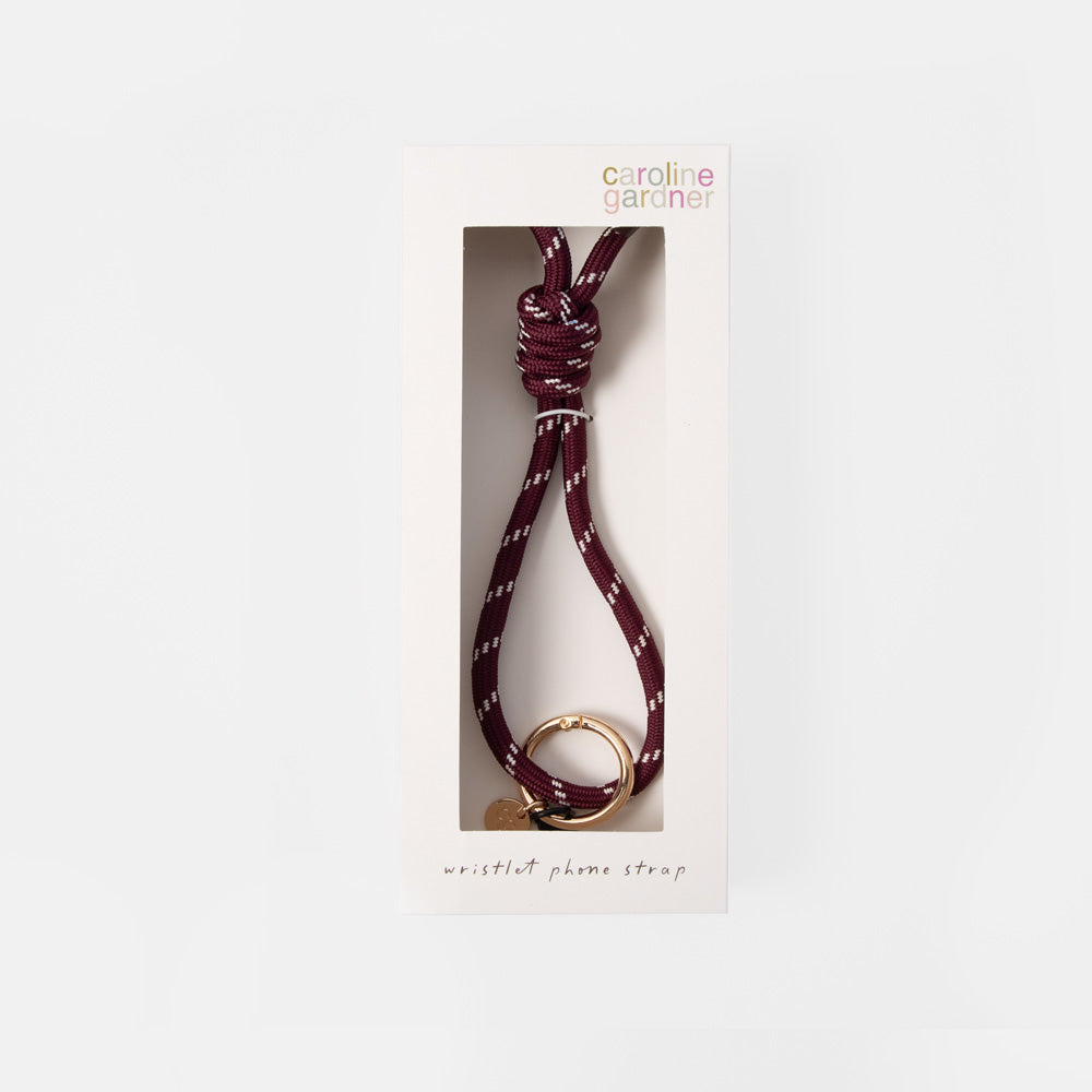A Carolina Gardner wristlet phone strap in burgundy with white dotted accents and a gold metal ring, shown inside its white retail packaging window box against a light gray background.