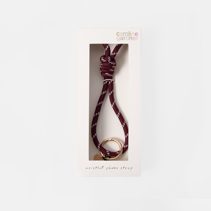 A Carolina Gardner wristlet phone strap in burgundy with white dotted accents and a gold metal ring, shown inside its white retail packaging window box against a light gray background.
