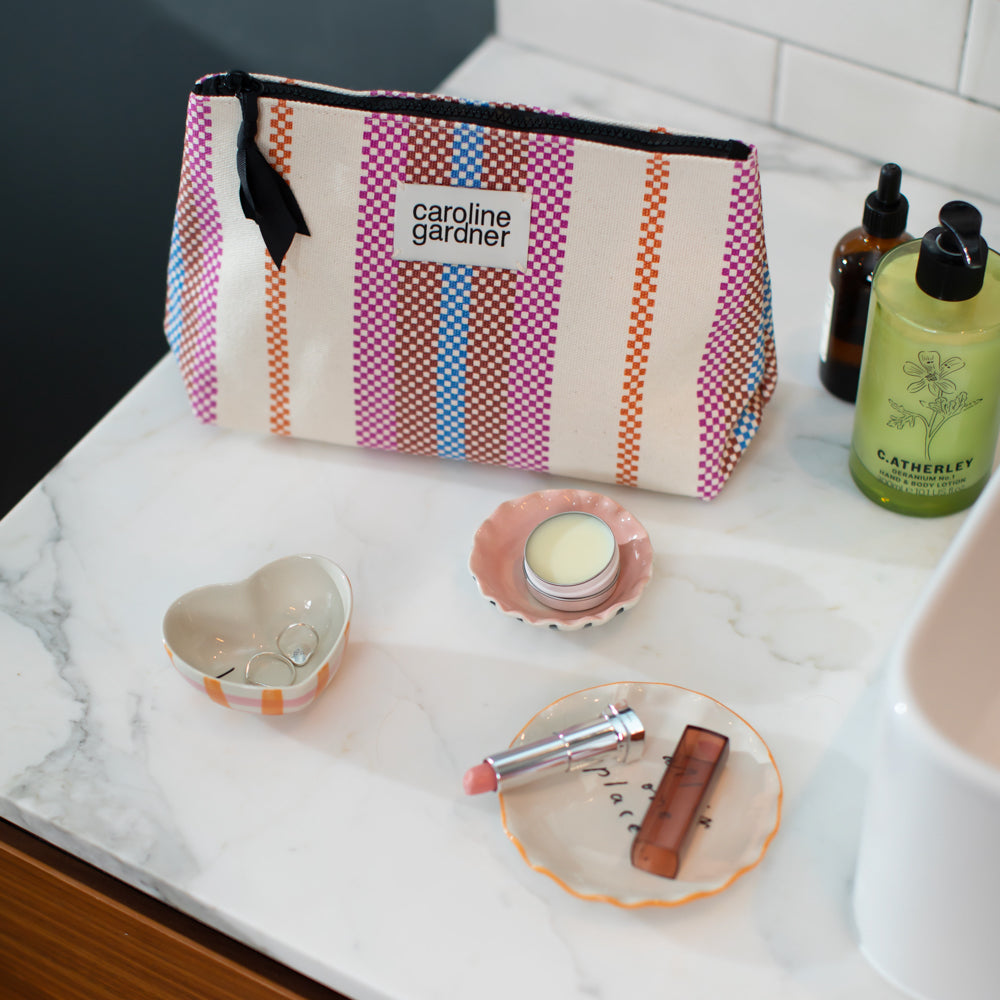 multi coloured checkerboard stripe beauty bag on a marble surface with hand cream, lipstick and trinket dishes