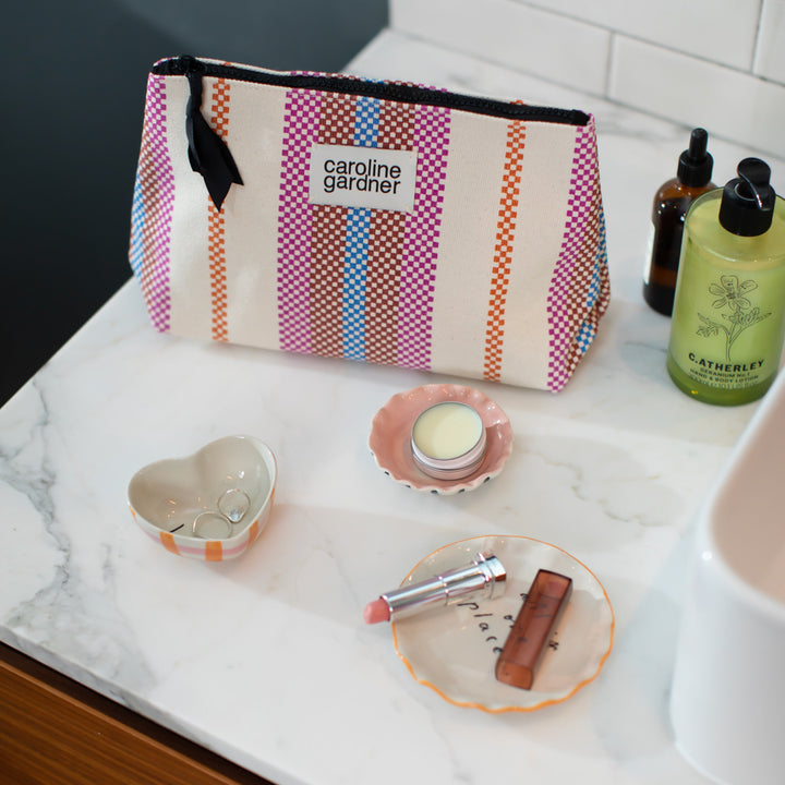 multi coloured checkerboard stripe beauty bag on a marble surface with hand cream, lipstick and trinket dishes