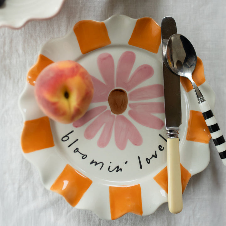 orange & white striped scalloped edge plate with a pink flower design and the words bloomin' lovely, with cutlery and a peach