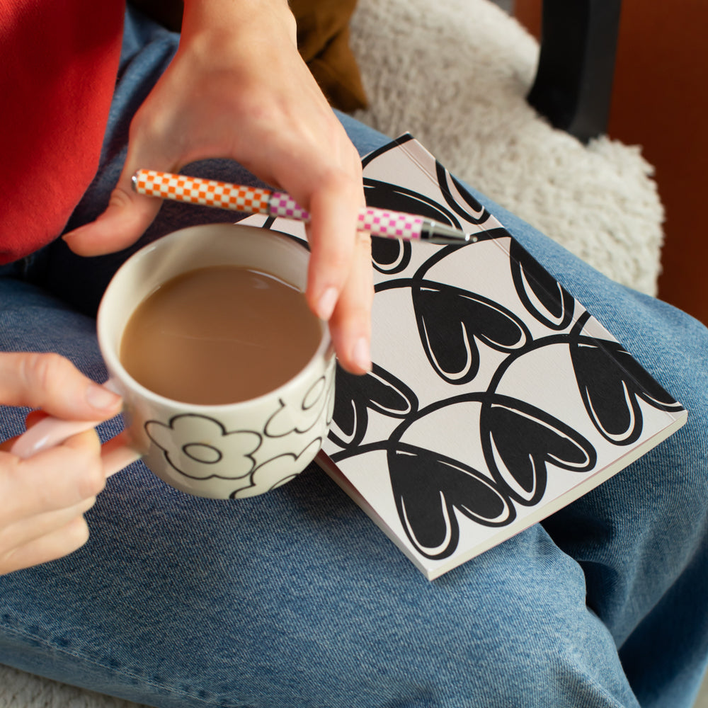 person holding a cup of tea in a flower print mug and a pen, with a black hearts print notebook on their lap