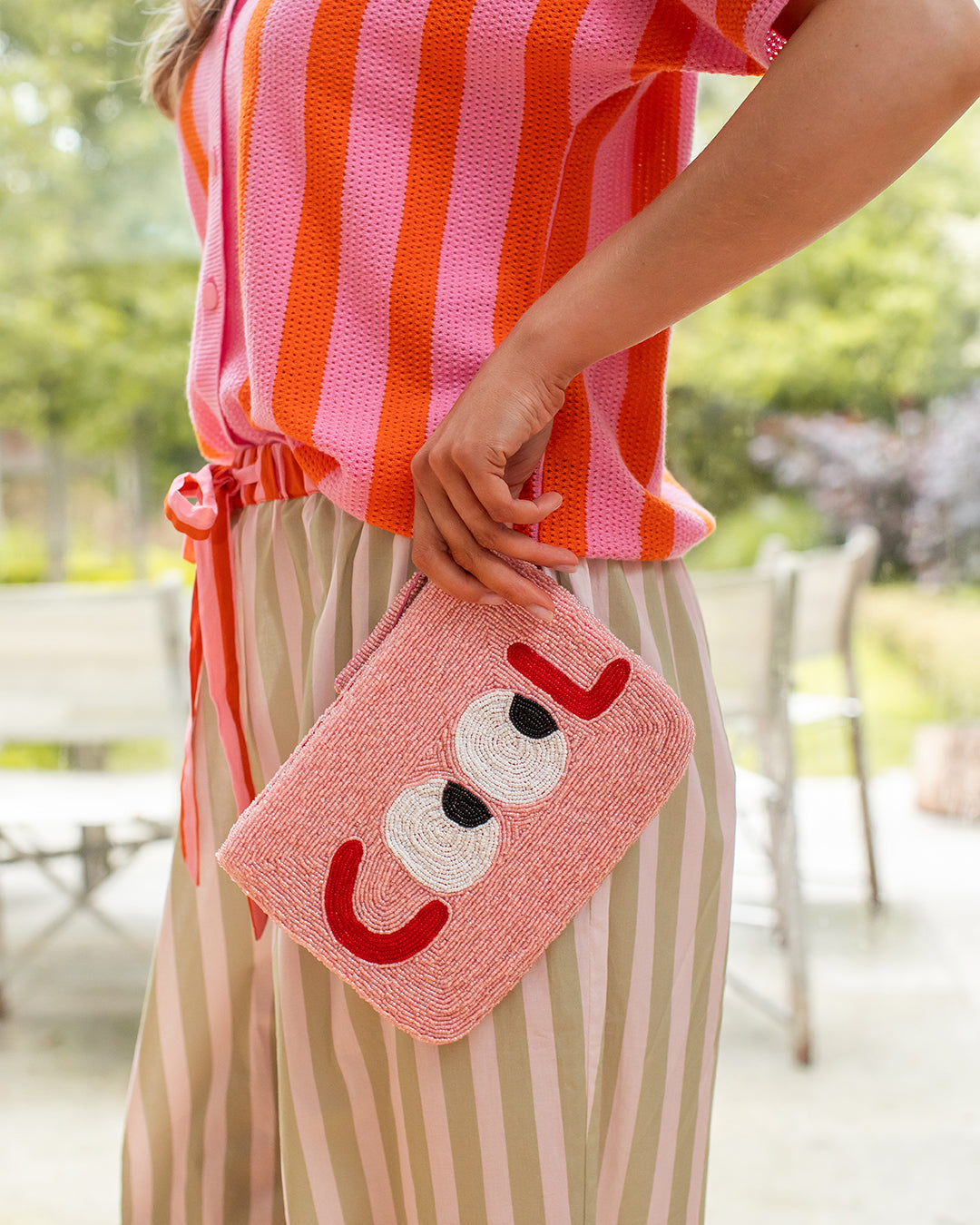 woman dressed in pink and red striped top with green and light pink striped trousers, holding pink clutch bag with the word COOL on it