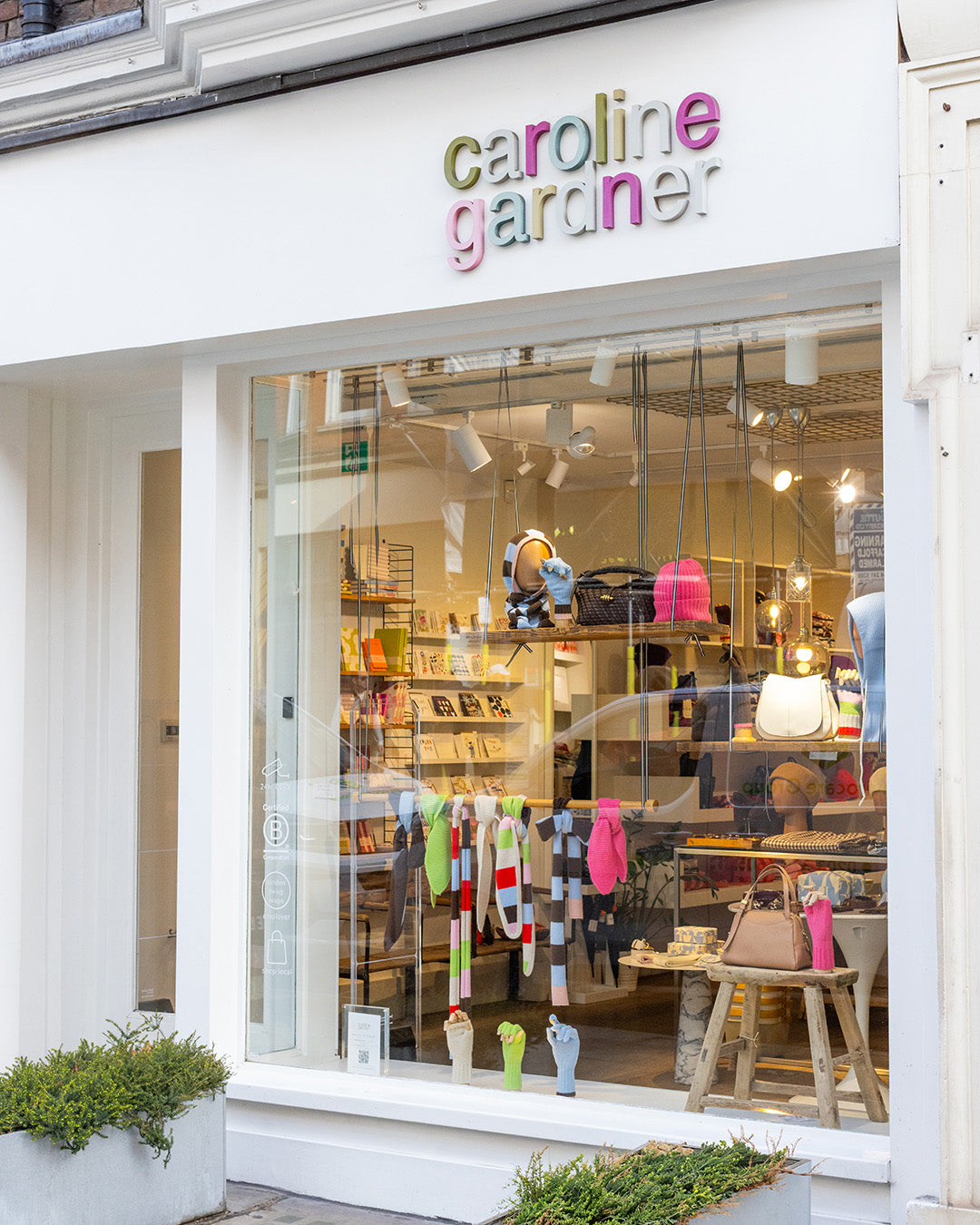Caroline Gardner boutique storefront with a large glass window displaying colorful knit garments, handbags, and interior decor, under a white facade with the brand name in multicolored letters.