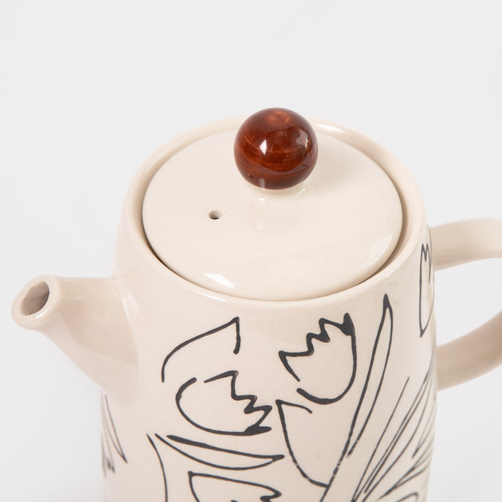 A close-up of a ceramic teapot with a cream-colored body, black line-art floral pattern, and a round brown lid knob, set against a plain light gray background.