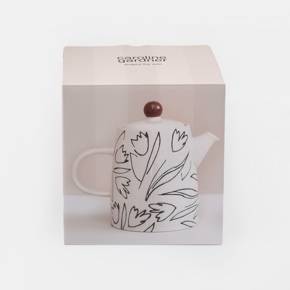 A white ceramic teapot with black line-art tulip patterns and a dark reddish-brown wooden lid knob, presented inside its light pink packaging box. The brand name 'caroline gardner' is visible on the top of the box.