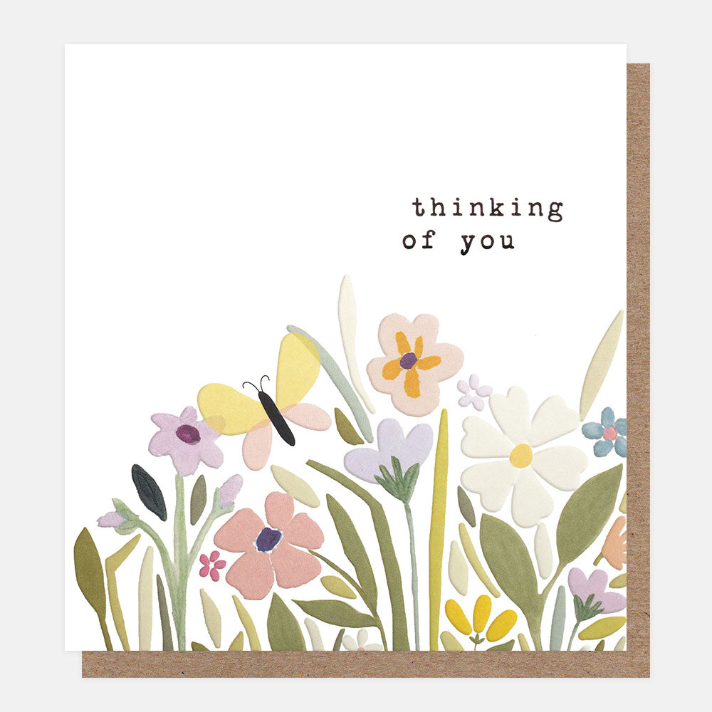 A white greetings card with a meadow of pink, lilac, white and yellow flowers and a yellow butterfly, with the text 'thinking of you' in black typewriter-style lettering, shown with a brown kraft envelope behind it.