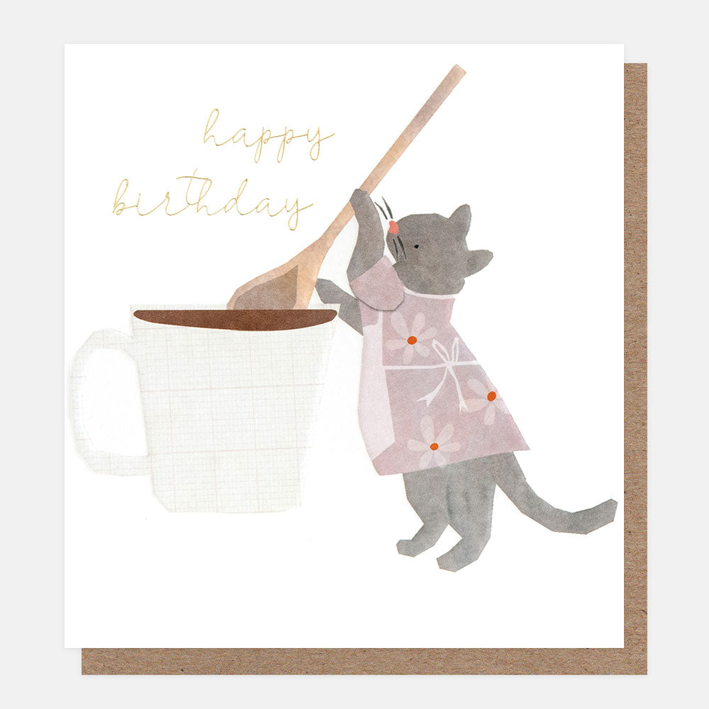 birthday card featuring a grey cat in a pink floral apron stirring a cup of tea with a wooden spoon and the text 'happy birthday' in gold