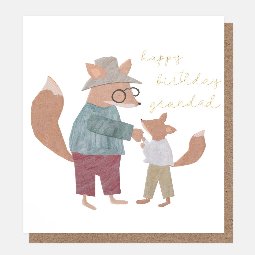 A white greetings card with a grandfather fox in a hat and glasses holding hands with a small fox and the text 'happy birthday grandad' in gold foil script, shown with a brown kraft envelope behind it.