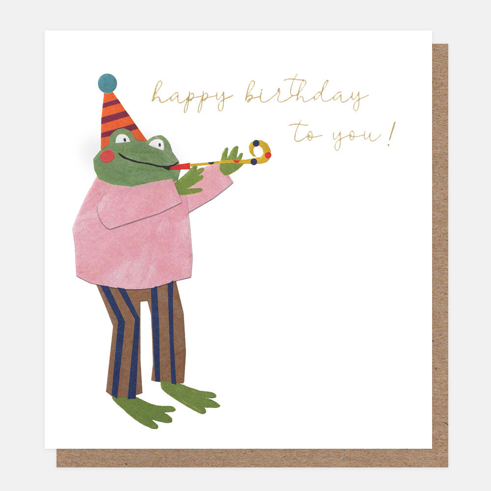 A white greetings card with a frog in a party hat and pink jumper blowing a party blower and the text 'happy birthday to you!' in gold foil script, shown with a brown kraft envelope behind it.