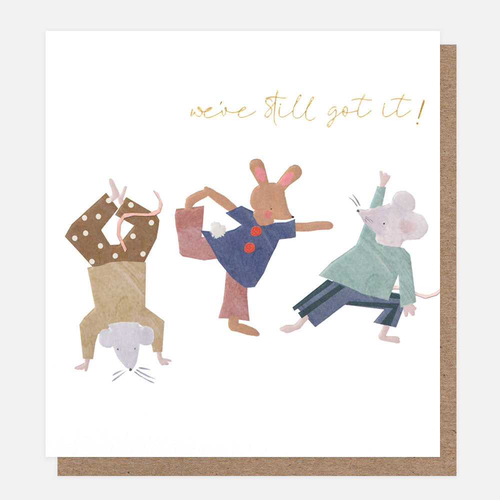 A white greetings card with two mice and a rabbit dancing, one doing a handstand, and the text 'we've still got it!' in gold foil script, shown with a brown kraft envelope behind it.