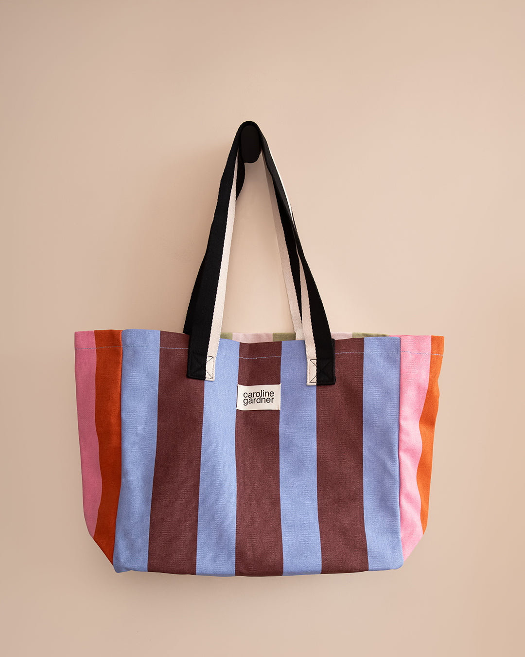 A striped canvas tote bag with black and white shoulder straps, featuring vertical stripes in pink, rust orange, light blue, and dark brown. A small white label on the front reads 'carolines dreamer'.