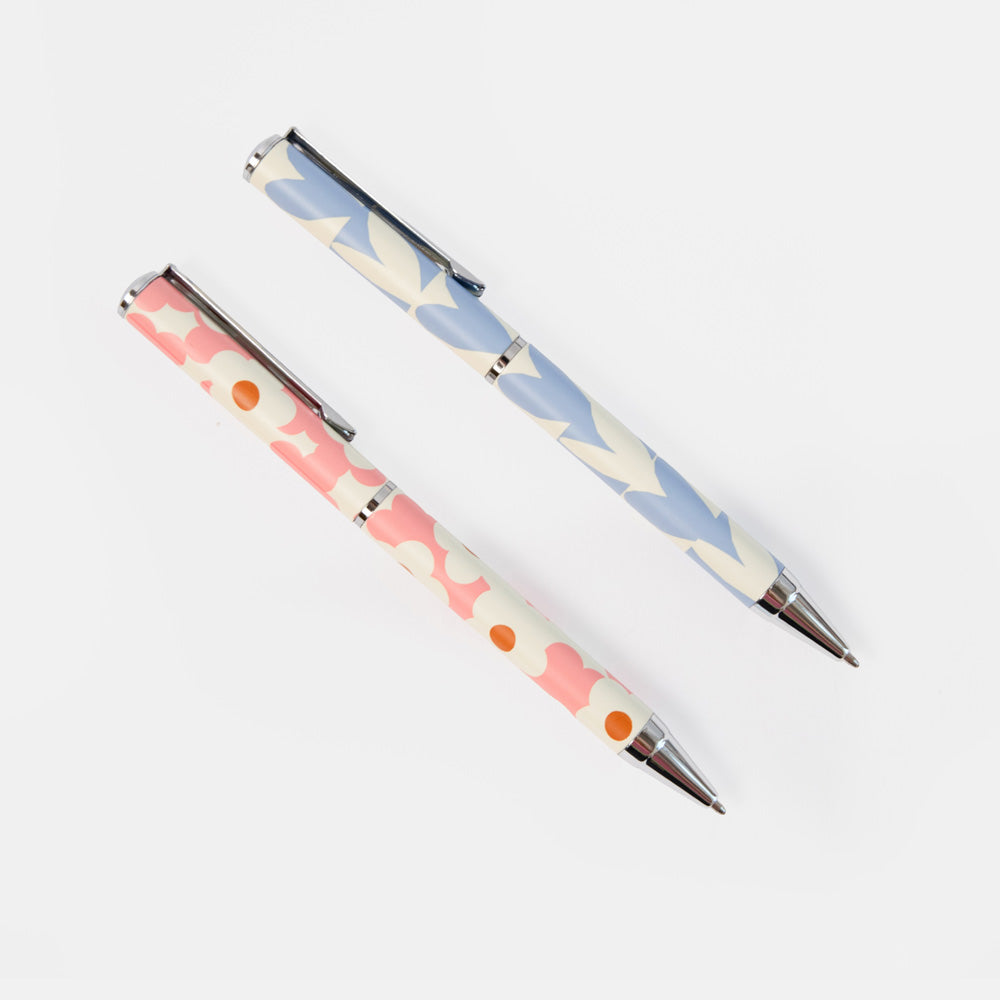 Two decorative ballpoint pens with a retro daisy pattern, one in pink and the other in light blue, displayed diagonally on a clean white background.