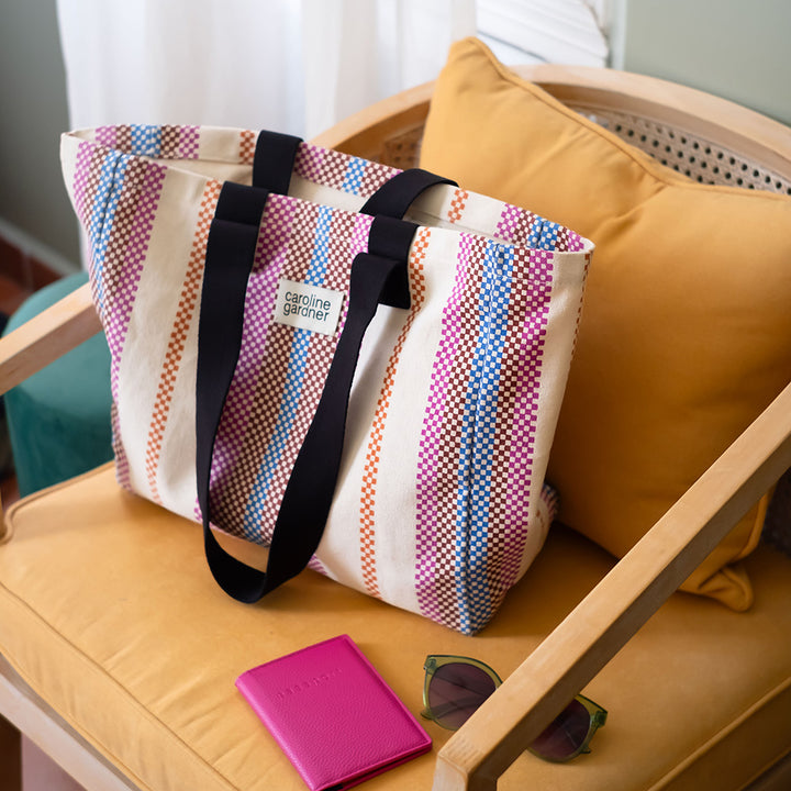 A striped canvas tote bag with black shoulder straps sits on a yellow chair next to a pink notebook and sunglasses.