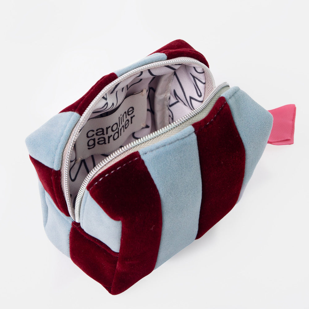 A striped cosmetic pouch with alternating light blue and deep burgundy velvet panels, shown open to reveal a white interior with black line art and the brand name 'caroline gardner'.