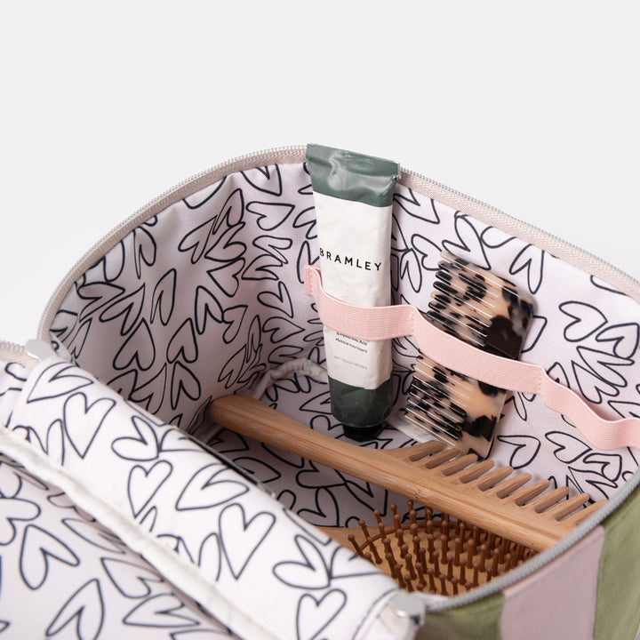 An open zippered vanity case with a white interior printed with black heart outlines and a pink elastic organizer. Visible items inside include a tube labeled Bramley, a leopard-print hairbrush, a wooden comb, and a brown bristle brush.